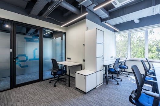 Desks with armchairs placed in spacious open space office with glass doors and white cupboards near big window in business center